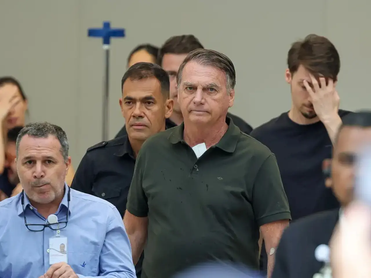 Rosto do ex-presidente Jair Bolsonaro caminhando. Ele aparece de camisa gola polo verde escura em destaque em meio a vários homens que o cercam