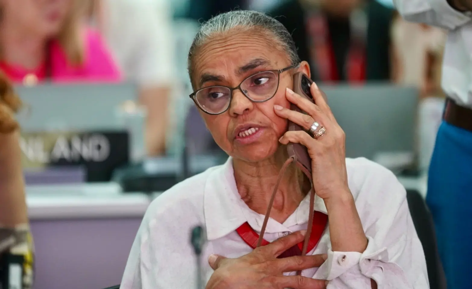 Marina Silva é uma mulher idosa, negra e magra, de cabelos grisalhos amarrados em um rabo de cavalo. Na foto, ela, que usa óculos de grau e blusa branca, está ao telefone.