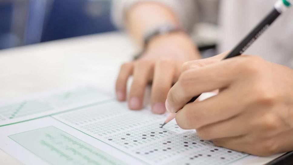 Mão de estudante preenchendo cartão-resposta de prova objetiva com lápis, marcando alternativas em gabarito de múltipla escolha.