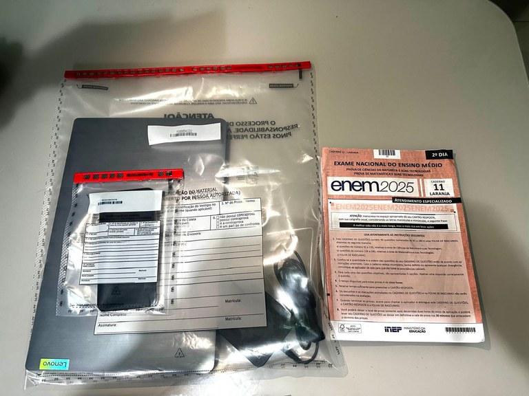 Sobre uma superfície clara, há dois itens principais: à esquerda, um pacote plástico transparente lacrado com tarja vermelha, contendo um notebook prateado da marca Lenovo, um carregador e um envelope menor com etiquetas de identificação; à direita, um caderno de provas do Exame Nacional do Ensino Médio (ENEM) 2025, identificado como “2º dia” e cor laranja, com instruções impressas na capa e o logotipo do INEP.