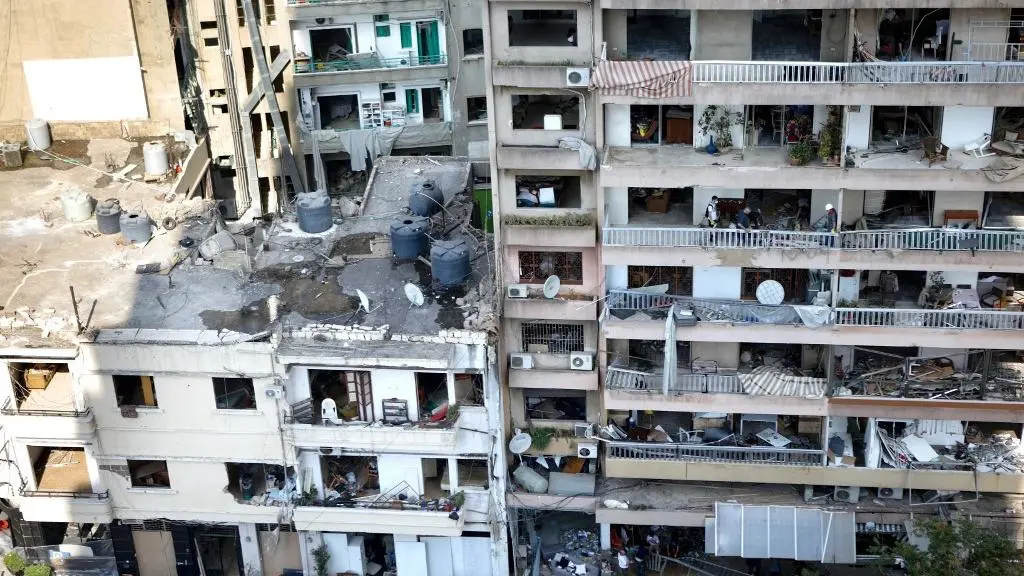 Na imagem, vista aérea de edifícios residenciais em uma área urbana, mostrando danos extensos, provavelmente após uma explosão. Os apartamentos têm varandas, muitas das quais estão cheias de destroços, janelas quebradas, estores danificados e pedaços de concreto. No telhado do edifício à esquerda, há grandes tanques de água e escombros espalhados. As fachadas dos edifícios mostram sinais visíveis de destruição.
