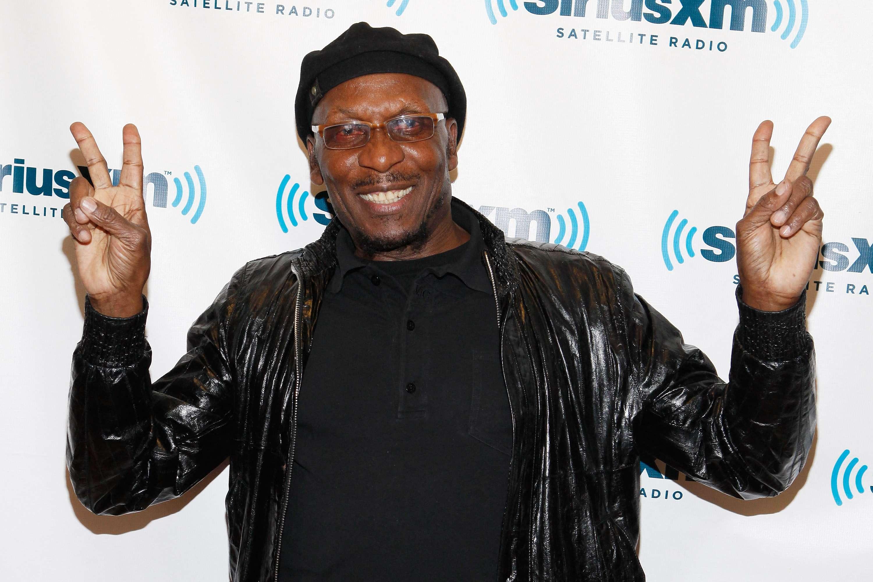 Imagem do lendário músico Jimmy Cliff sorrindo e fazendo o sinal de paz com as duas mãos, usando um boné e jaqueta de couro preta.