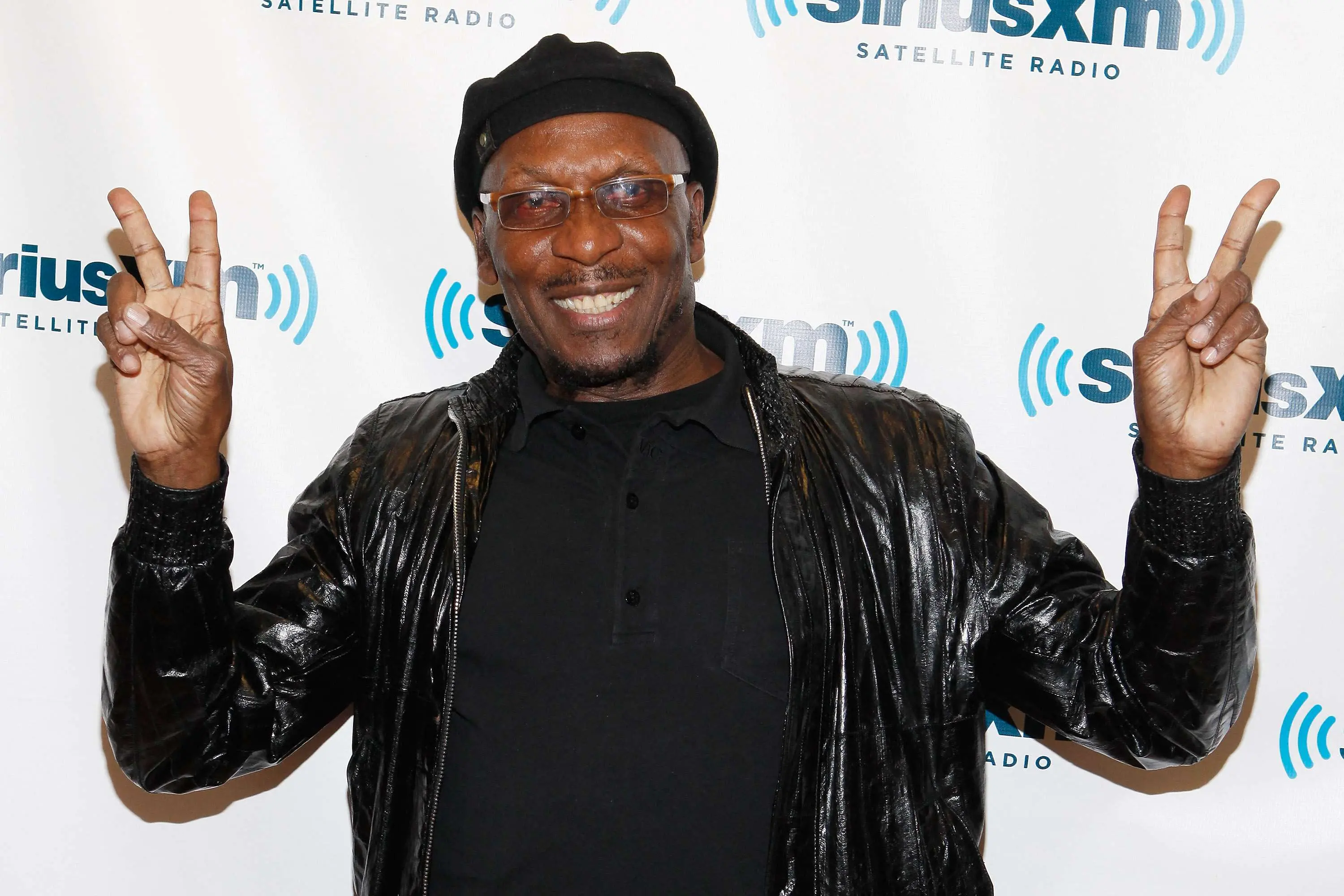Imagem do lendário músico Jimmy Cliff sorrindo e fazendo o sinal de paz com as duas mãos, usando um boné e jaqueta de couro preta.