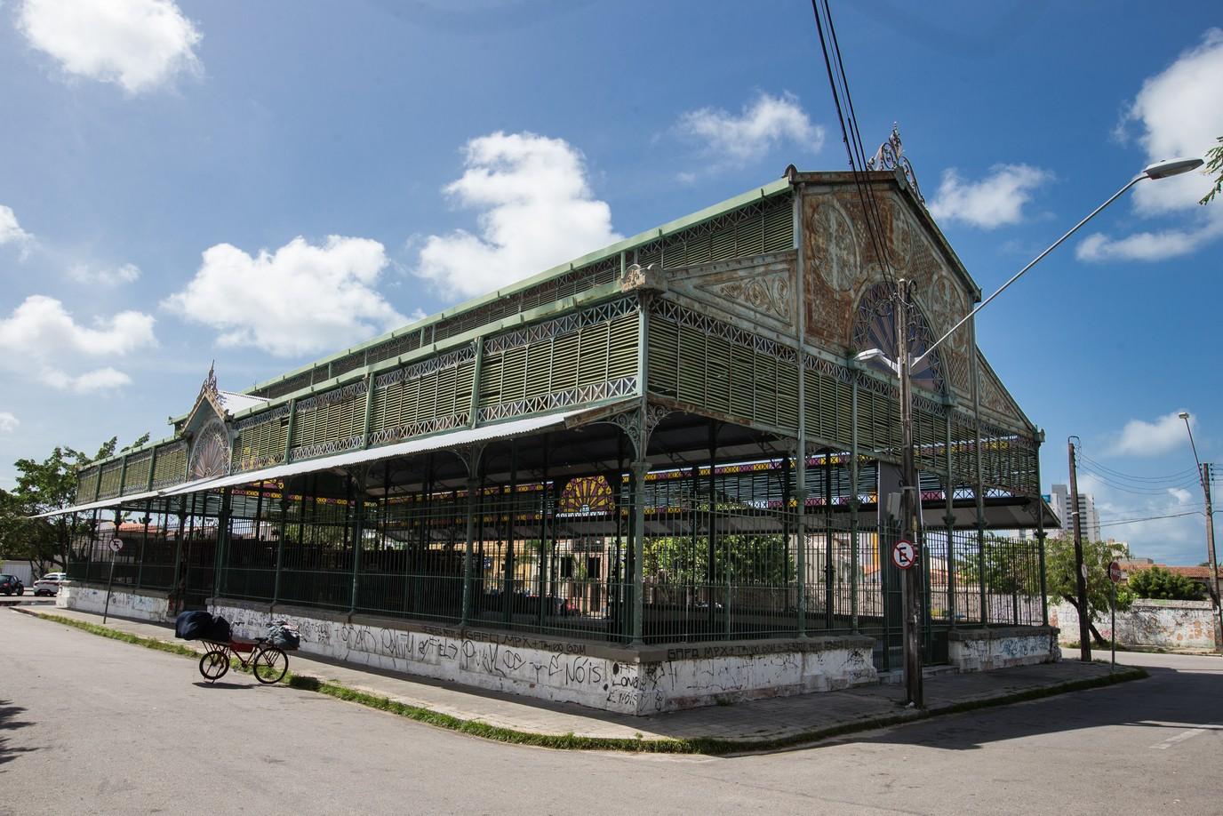 Uma foto externa do Mercado dos Pinhões, em Fortaleza. É uma estrutura metálica histórica (ferro fundido) com teto e paredes laterais em venezianas de metal pintadas de verde, sob um céu azul claro com nuvens. Uma carroça de bicicleta está estacionada na rua lateral no canto inferior esquerdo.