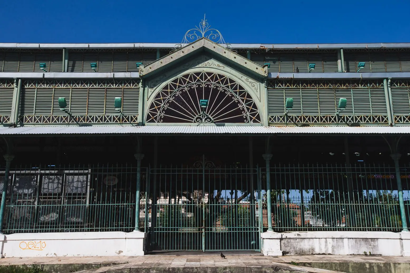 Um plano frontal e mais próximo da entrada principal de uma estrutura de mercado, identificada como Mercado da Aerolândia. A arquitetura é semelhante à do Mercado dos Pinhões, com painéis de metal verde e um arco ornamentado com detalhe de vitral ou tela de ferro na fachada superior. O portão está fechado.