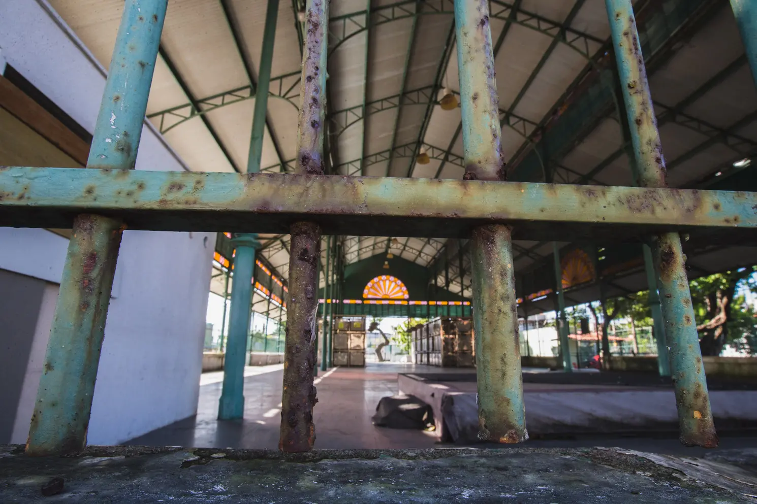 Um close-up através das barras de metal de uma cerca ou portão do Mercado da Aerolândia, mostrando sinais visíveis de desgaste e ferrugem nas barras pintadas de verde/azul. O fundo, em foco suave, mostra o interior coberto da estrutura do mercado.