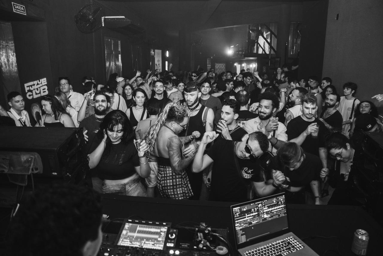 Festa em clube noturno com pista de dança cheia, pessoas aproveitando música eletrônica, DJ tocando em mesa com laptop e equipamentos, iluminação baixa e ambiente vibrante.