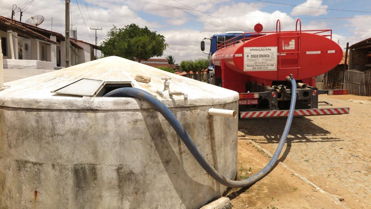 Imagem mostra um caminhão-pipa vermelho estacionado em uma rua de calçamento, abastecendo uma grande cisterna de concreto por meio de uma mangueira azul conectada à abertura superior do reservatório. Casas simples aparecem ao fundo, junto a postes e fiação elétrica, em um cenário de céu parcialmente nublado. A cena retrata o abastecimento de água em uma comunidade.