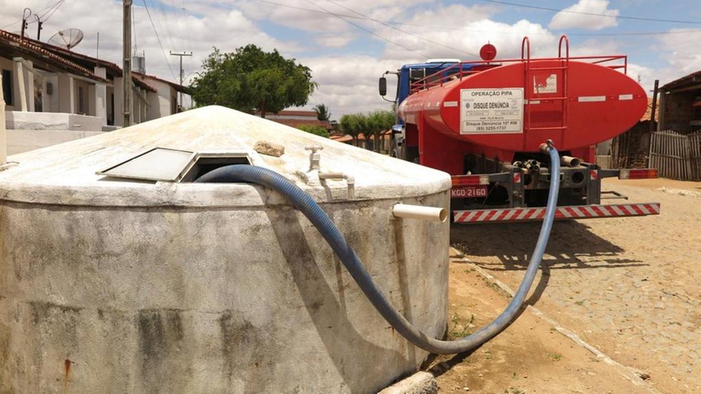 Imagem mostra um caminhão-pipa vermelho estacionado em uma rua de calçamento, abastecendo uma grande cisterna de concreto por meio de uma mangueira azul conectada à abertura superior do reservatório. Casas simples aparecem ao fundo, junto a postes e fiação elétrica, em um cenário de céu parcialmente nublado. A cena retrata o abastecimento de água em uma comunidade.