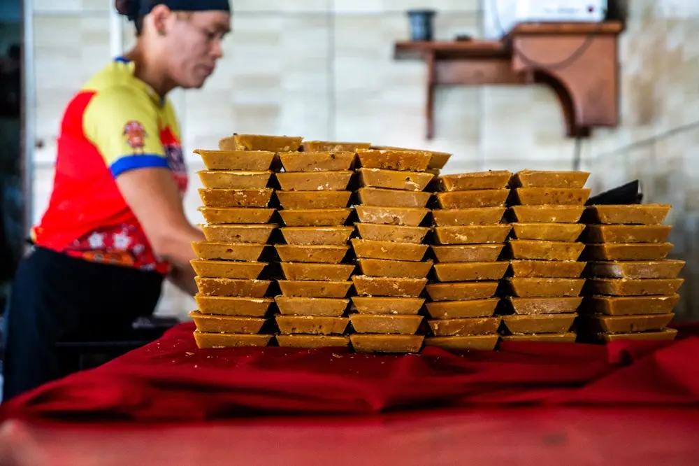 Na imagem, pilhas de rapadura em formato de tijolo de cor âmbar dourado estão empilhadas sobre uma toalha vermelha escura. No fundo, fora de foco, uma pessoa vestindo uma camiseta vermelha e amarela e um avental escuro está de pé em uma sala de processamento ou loja.