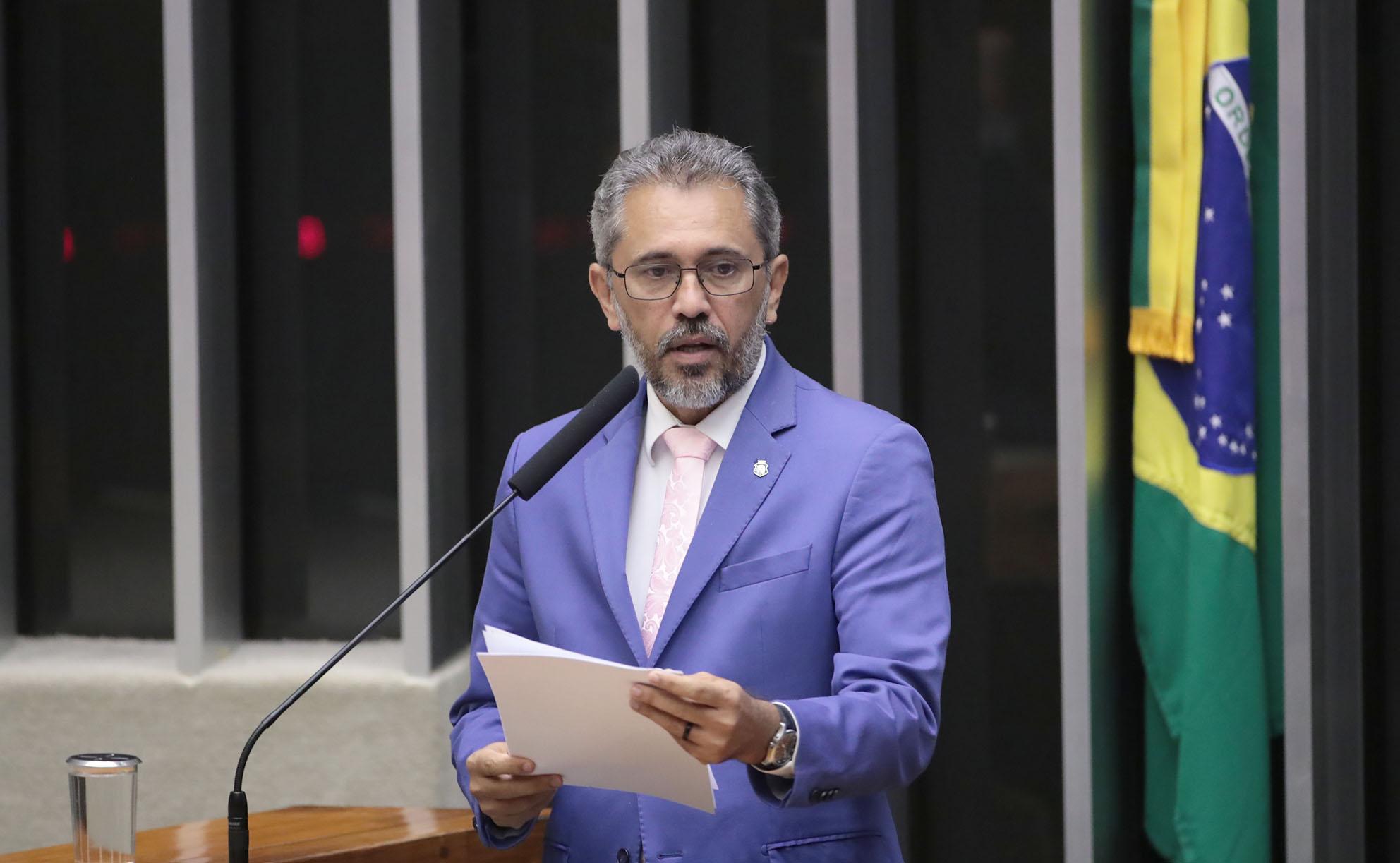 Homem de meia-idade, de barba grisalha curta e óculos, veste terno azul com camisa branca e gravata rosa clara. Trata-se de Elmano de Freitas. Ele aparece em um púlpito, segurando folhas de papel enquanto fala ao microfone. Ao fundo, há barras verticais escuras e parte de uma bandeira do Brasil visível à direita da imagem.