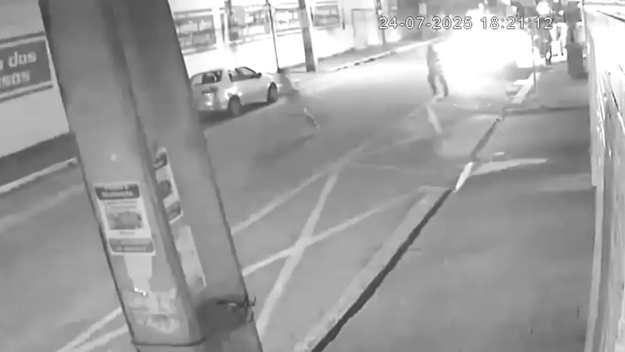 A imagem de uma câmera de segurança mostra um momento em que um preso tentou fugir algemado e foi baleado por policial militar, em Fortaleza. A imagem está em preto e branco, há um poste em primeiro plano e carros na rua.