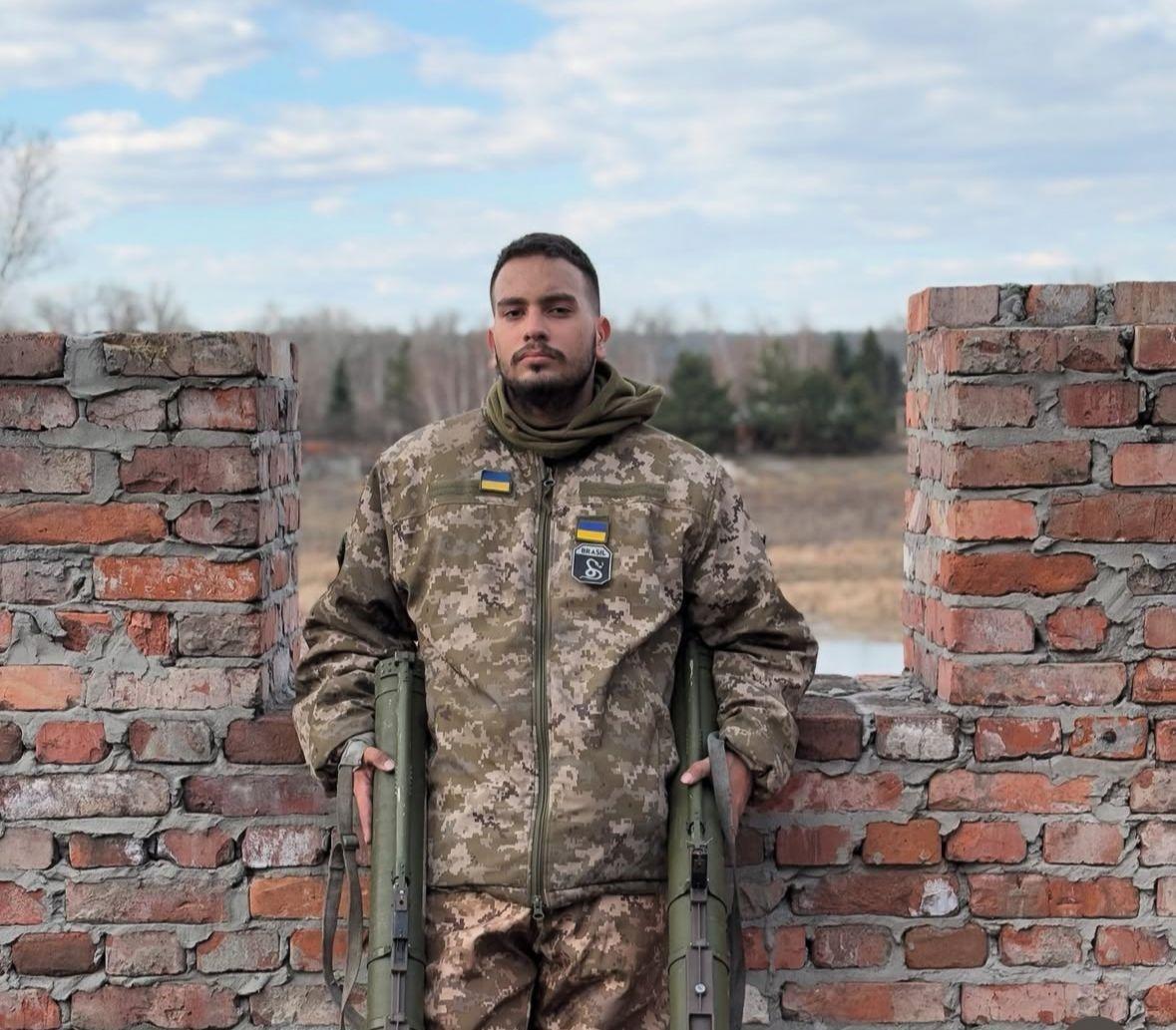 Soldado aparece em pé, vestido com uniforme camuflado do Exército ucraniano, apoiado entre duas paredes de tijolos.