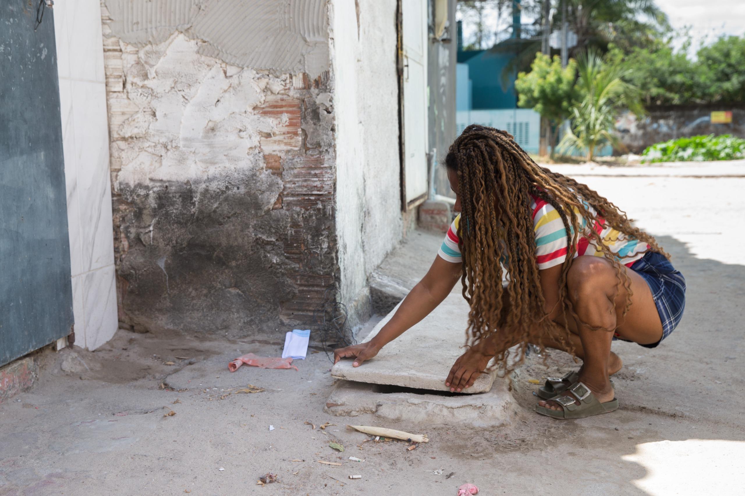 Mulher de cabelos trançados, camiseta listrada e shorts jeans agachada em uma calçada de concreto, abrindo tampa de bueiro em rua.