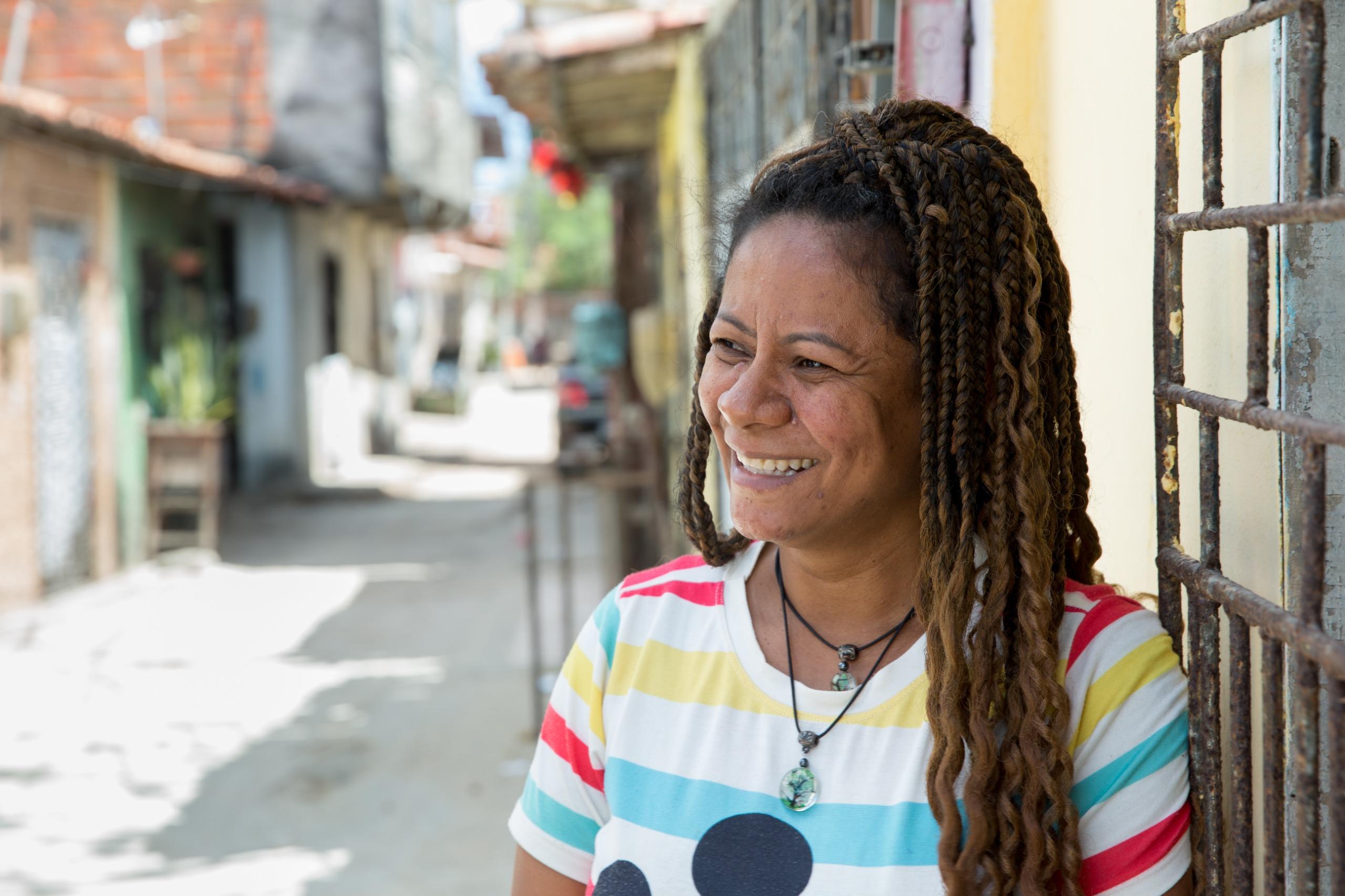 Mulher sorrindo com tranças longas e camiseta colorida em uma rua de bairro.
