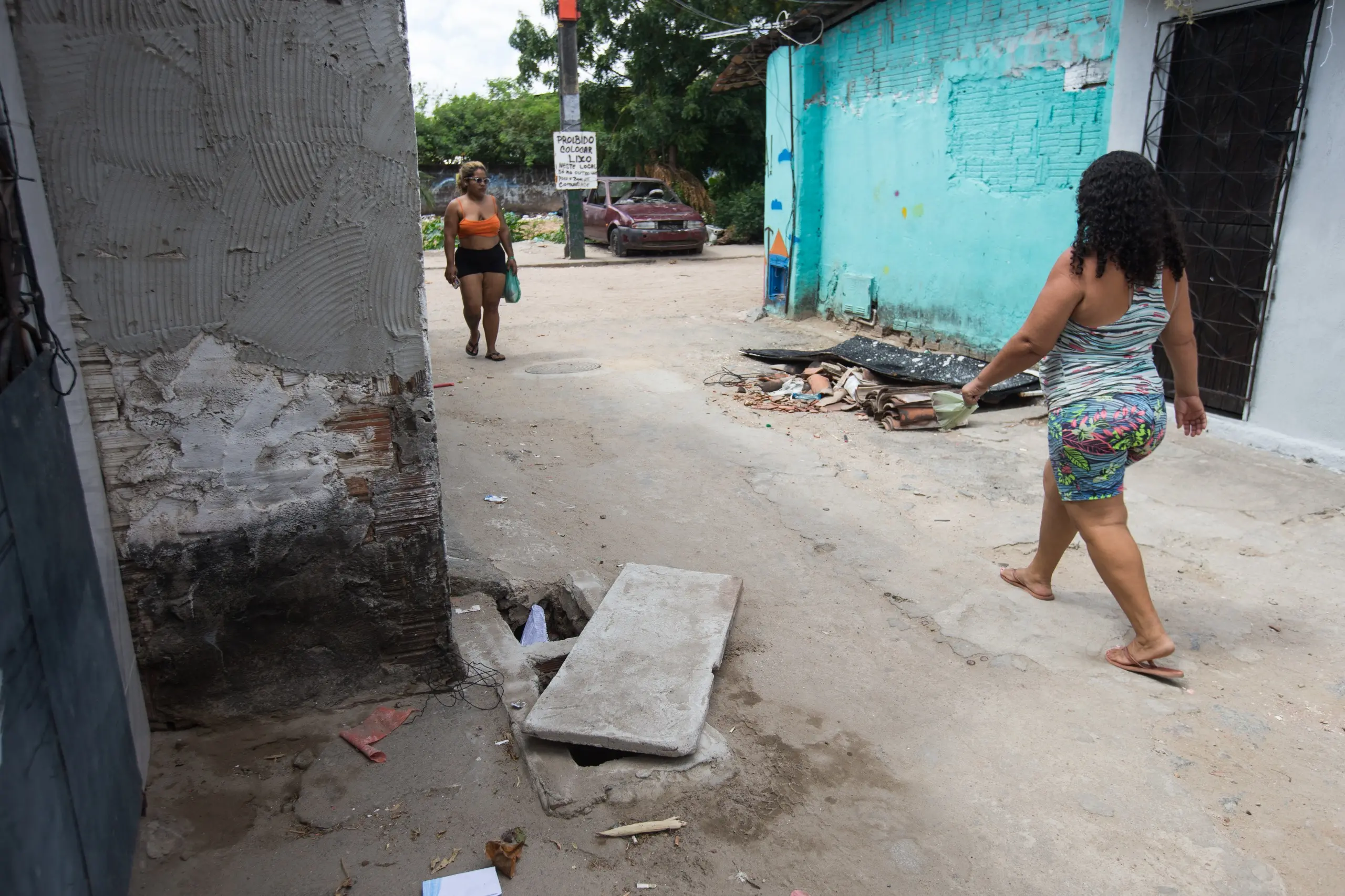 Duas mulheres caminham por uma rua irregular de bairro, com casas simples, entulho e um carro ao fundo.
