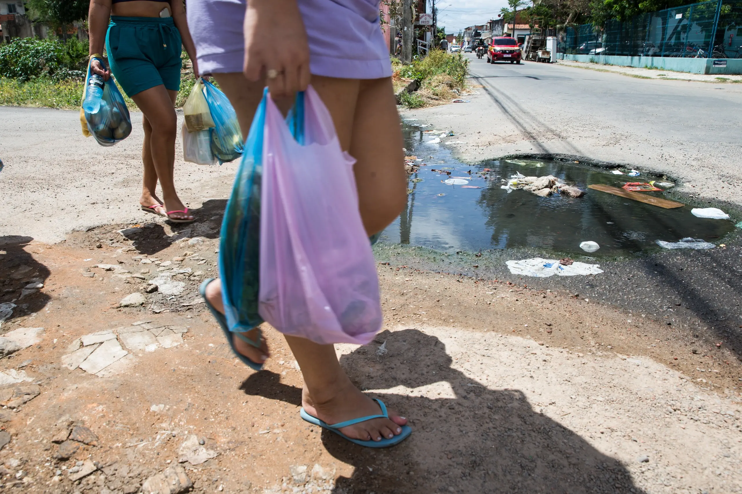 Mulheres com sacolas de compras na mão cruzam poça de esgoto na rua.