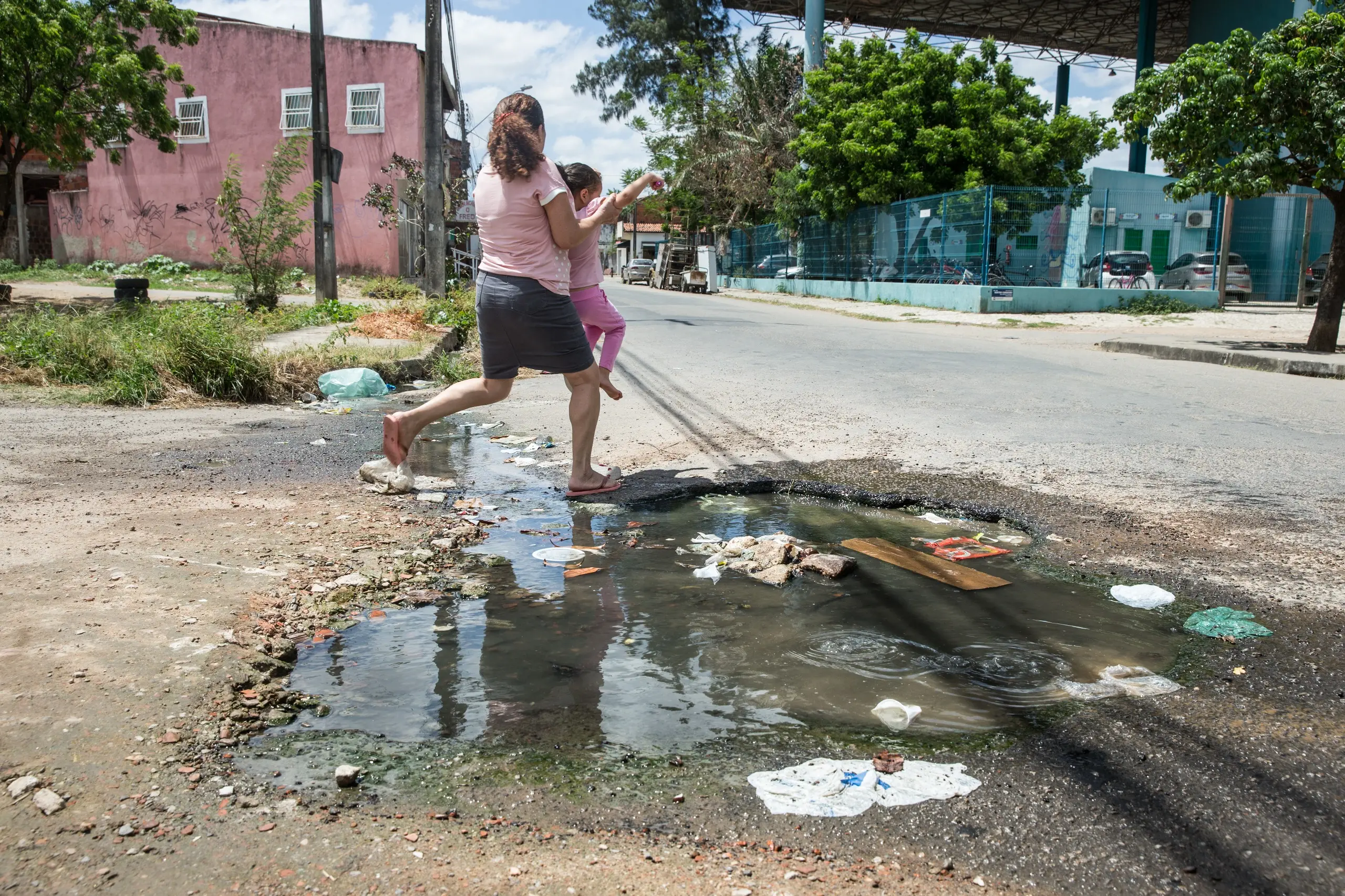 Uma mulher descalça carrega uma criança pequena nos braços enquanto pula sobre uma grande poça de água suja e lixo em uma rua de terra.
