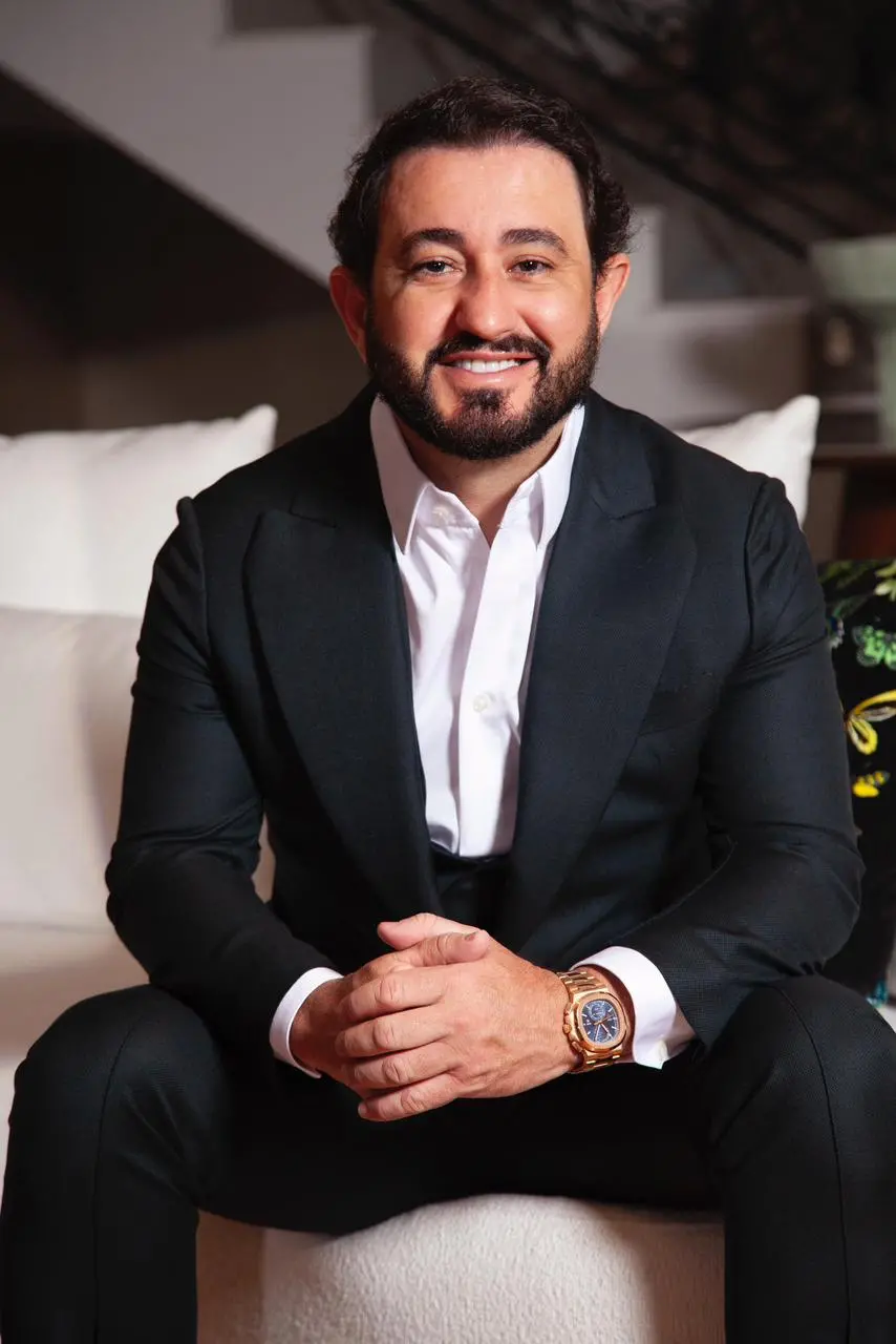 Imagem mostra homem branco e com barba. Usa terno e sorri para a foto.