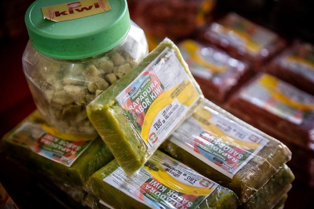 Na imagem, uma foto em close-up, com profundidade de campo rasa, mostra vários pedaços de rapadura de cor verde, embalados individualmente em plástico transparente, empilhados sobre uma superfície. Um rótulo parcialmente visível nas embalagens diz 