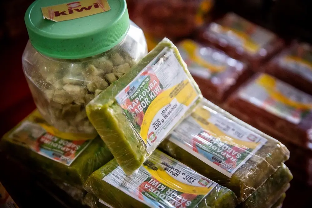 Na imagem, uma foto em close-up, com profundidade de campo rasa, mostra vários pedaços de rapadura de cor verde, embalados individualmente em plástico transparente, empilhados sobre uma superfície. Um rótulo parcialmente visível nas embalagens diz 