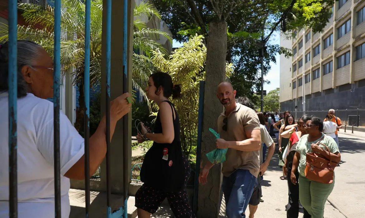 Imagem mostra participantes da PND 2025 acessam um local de aplicação da prova através de um portão, com várias pessoas em primeiro plano e uma longa fila se formando ao fundo da rua.