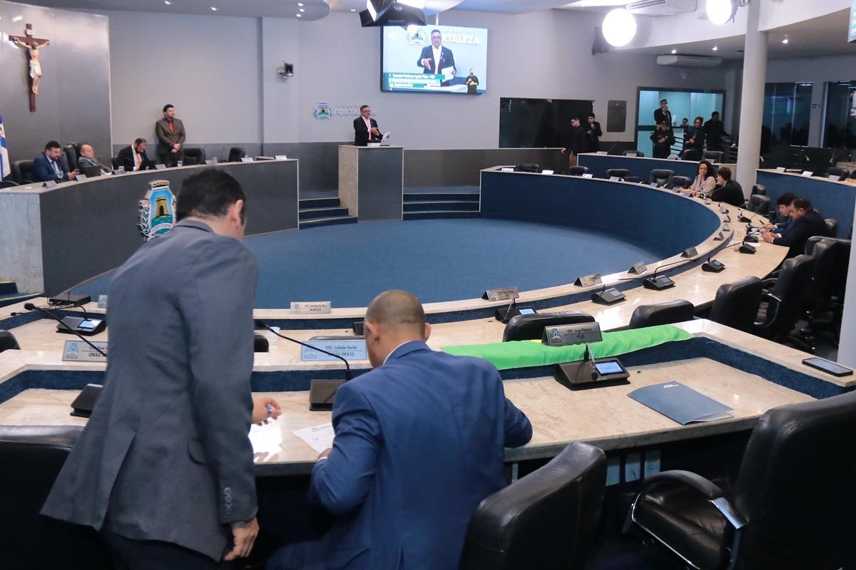Plenário da Câmara Municipal de Fortaleza, com vereadores reunidos em torno da mesa circular; alguns conversam e outros acompanham um orador que fala em púlpito ao fundo, diante de um telão.