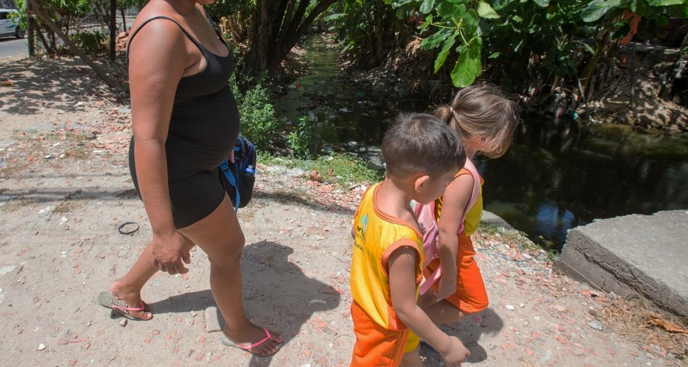 Mulher de vestido preto segurando uma mochila azul caminha atrás de duas crianças, uma menina e um menino, vestidos com a farda laranja e amarela da Prefeitura de Fortaleza. Eles estão sob o sol num caminho de terra. Ao fundo, há um córrego cheio de lixo cercado por vegetação.