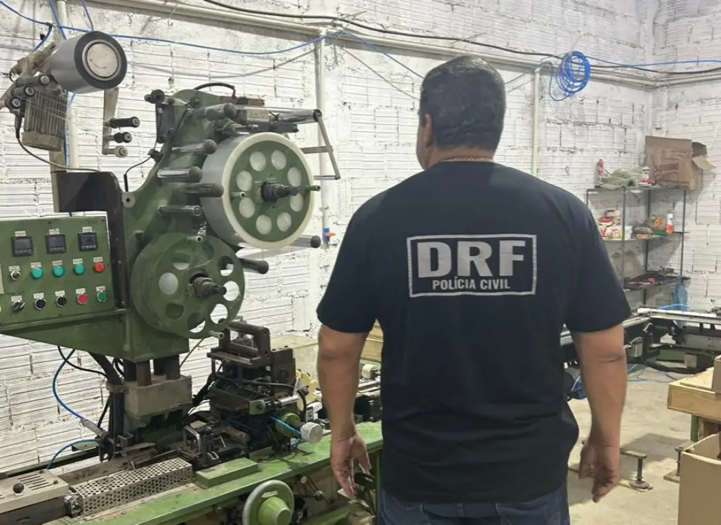 A imagem mostra um policial civil da DRF, de costas, em uma fábrica clandestina de cigarros, onde havia maquinário importado, no Interior do Ceará.