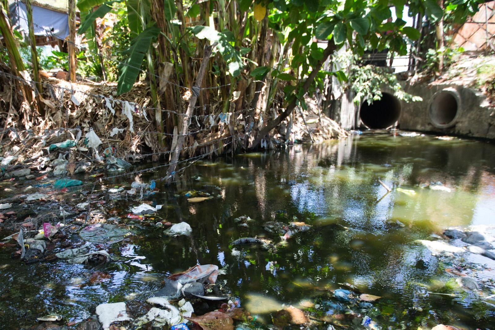 Uma cena que foca na poluição do Riacho Maceió. A água escura reflete a vegetação de bananeiras, e há uma grande quantidade de lixo acumulada ao longo das margens e flutuando na superfície. Ao fundo, tubos de esgoto/drenagem podem ser vistos.