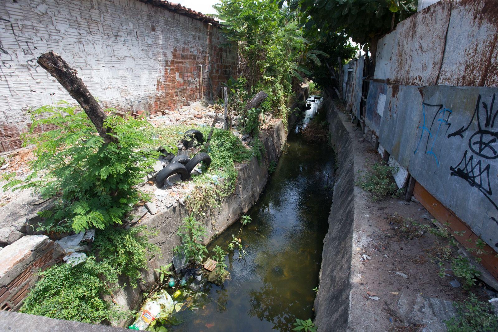 Uma vista aérea de um trecho canalizado (com paredes de concreto) do Riacho Maceió, que corre entre muros. A água está escura, e as margens apresentam crescimento de vegetação, lixo acumulado e alguns pneus descartados.