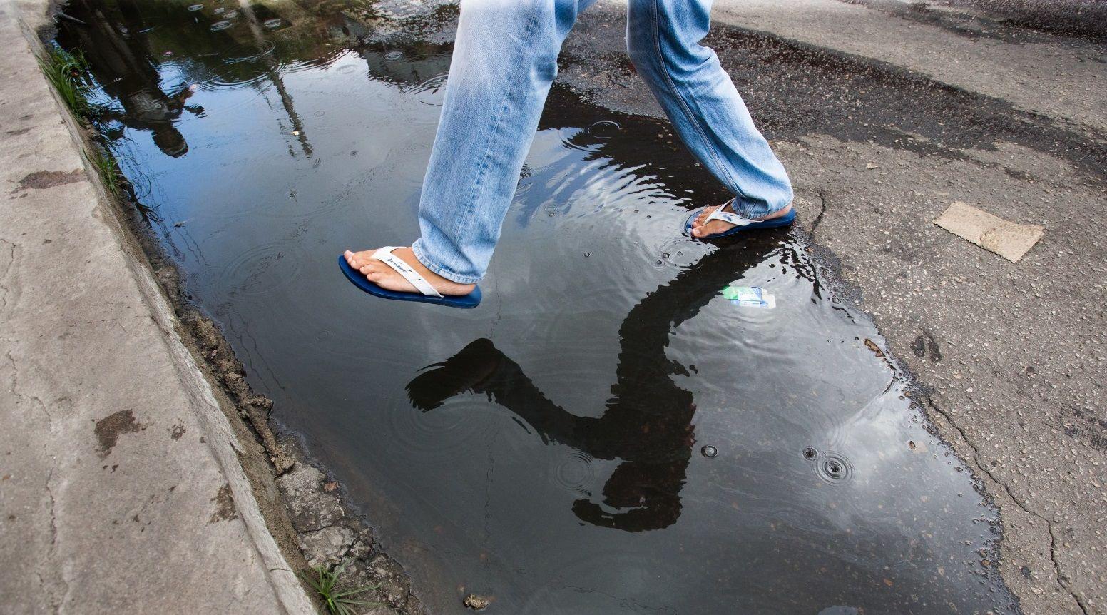 Um plano detalhado das pernas de uma pessoa de jeans e chinelos, pulando sobre uma poça longa e rasa de água escura/esgoto na sarjeta de uma rua. A silhueta refletida do corpo aparece na superfície da água.