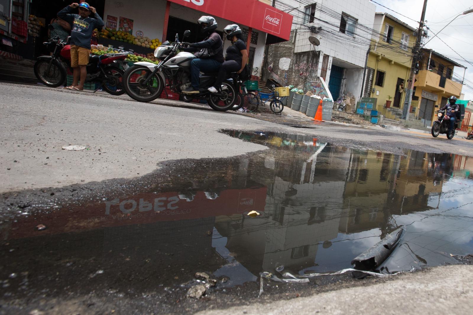 Uma rua no bairro Vicente Pinzón (Fortaleza), onde uma grande poça de água suja (esgoto a céu aberto) reflete os prédios e a loja ao redor. Pessoas em motocicletas e a pé transitam na via inclinada, ignorando o espelho d'água contaminado.