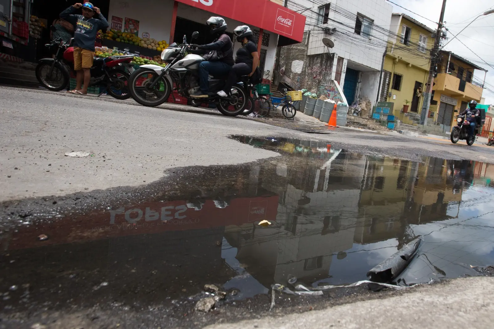 Uma rua no bairro Vicente Pinzón (Fortaleza), onde uma grande poça de água suja (esgoto a céu aberto) reflete os prédios e a loja ao redor. Pessoas em motocicletas e a pé transitam na via inclinada, ignorando o espelho d'água contaminado.