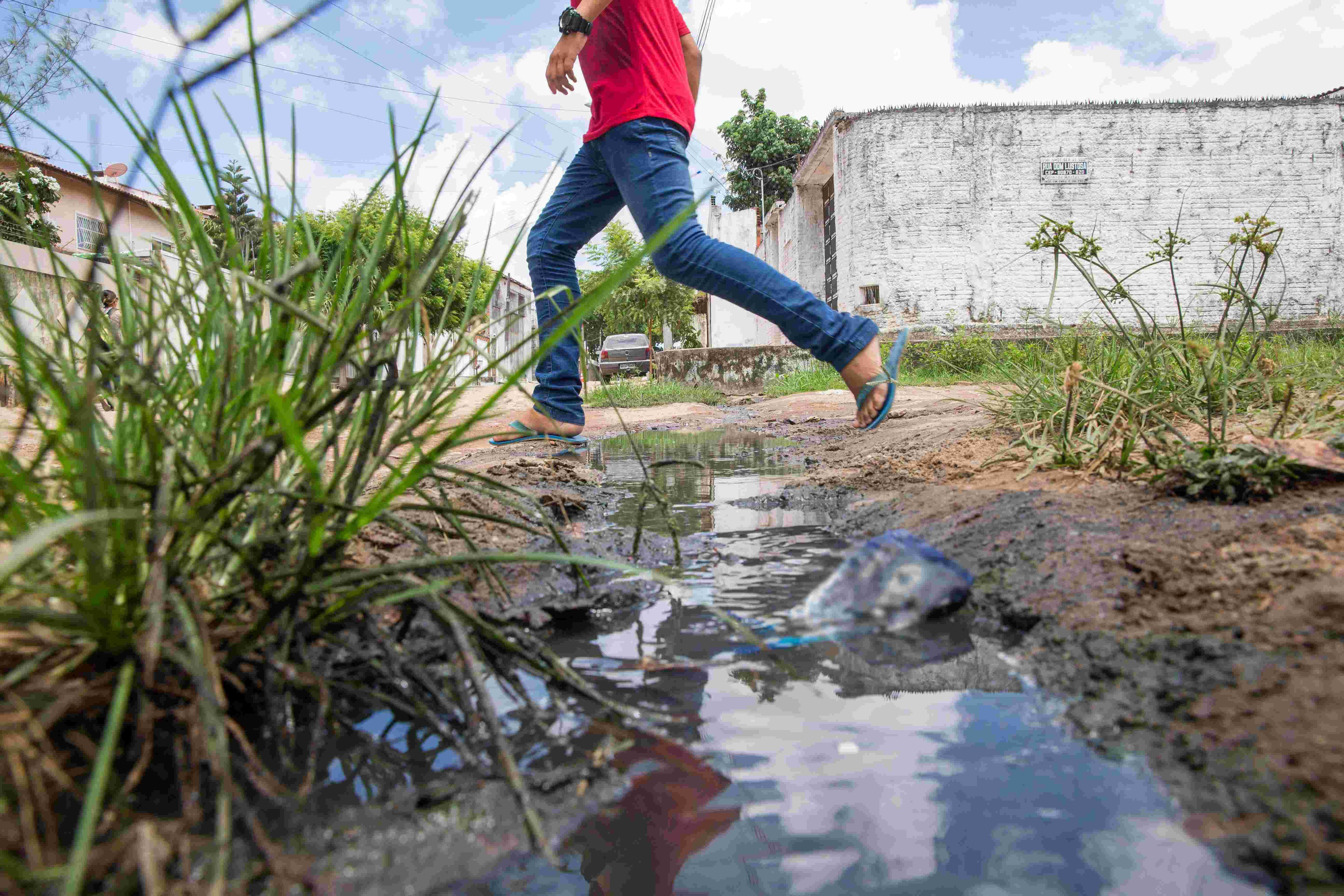 Uma perspectiva de baixo para cima, mostrando um homem de camisa vermelha e calça jeans pulando ou atravessando um pequeno córrego de água suja/esgoto que corre em uma vala de terra no meio do mato, em uma área residencial.