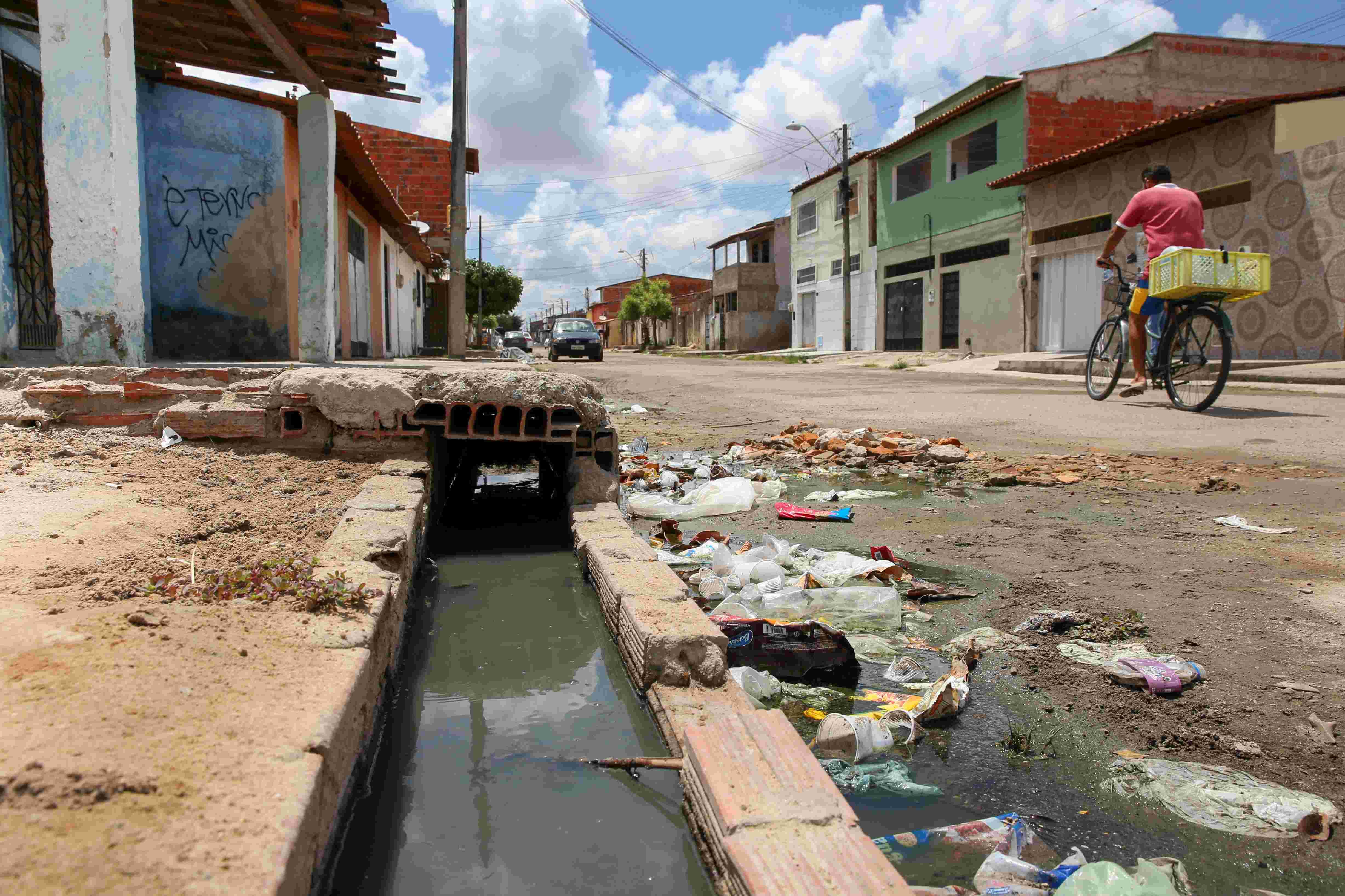 Uma vista de uma rua de bairro em Fortaleza com esgoto a céu aberto correndo em uma vala de alvenaria. Há uma grande quantidade de lixo (plástico, papel, etc.) acumulado na margem da vala. Ao fundo, casas simples e um ciclista transitam na rua.