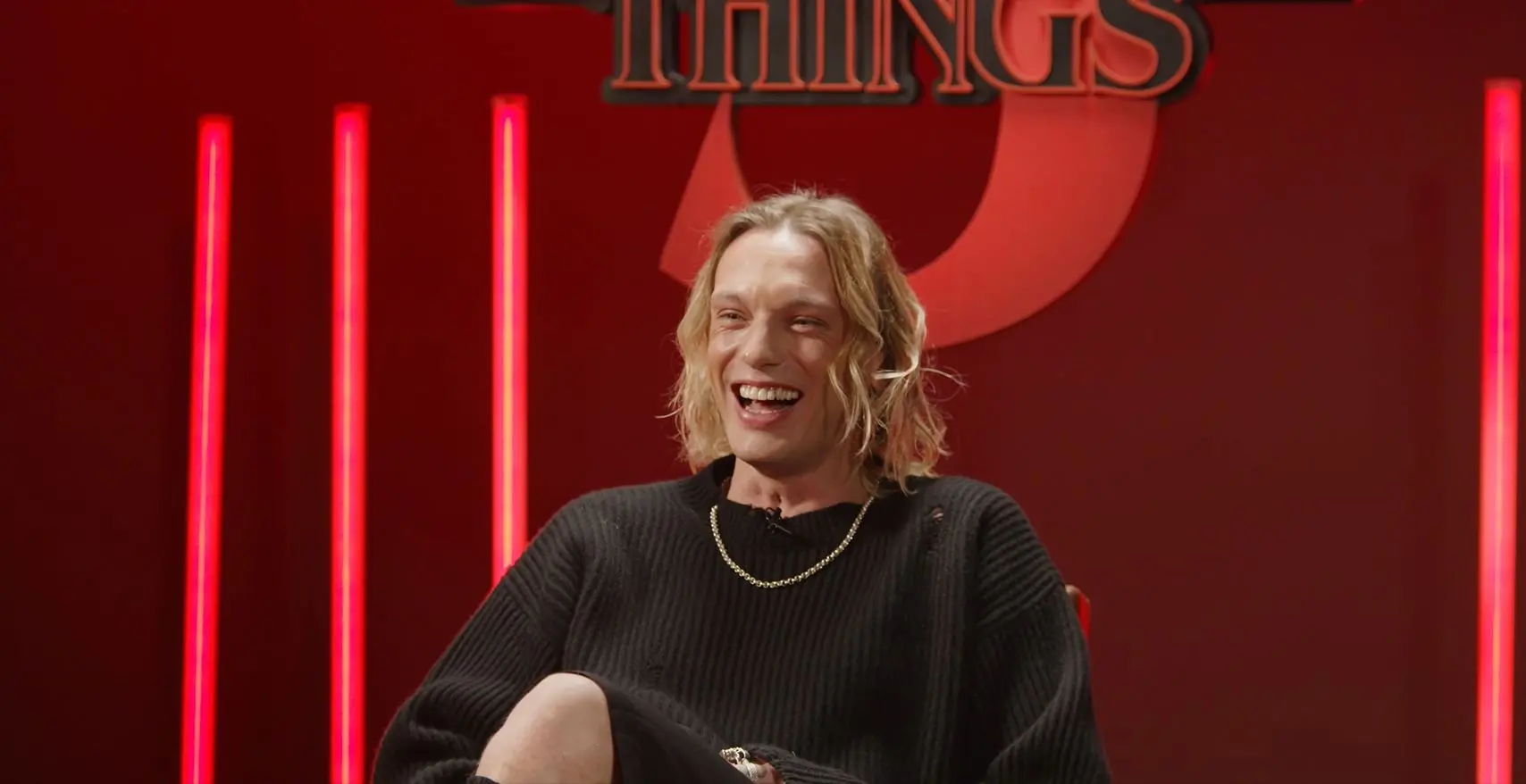 Jamie Campbell Bower com camisa preta, sorrindo, sentado em um cenário vermelho com alusão à Stranger Things.