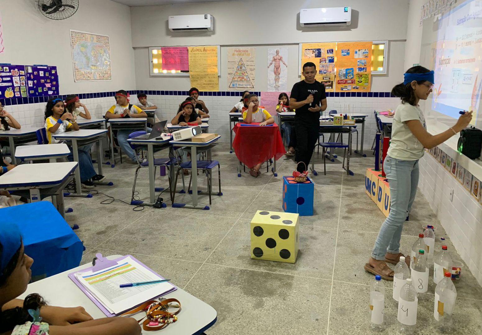 Sala de aula com alunos do ensino fundamental sentados em grupos, participando de uma atividade lúdica de matemática. No centro, há cubos grandes em forma de dado, garrafas plásticas numeradas no chão e um projetor apontado para o quadro. Uma aluna escreve no quadro enquanto usa uma venda azul, e um professor acompanha a dinâmica. Os cartazes coloridos nas paredes mostram conteúdos escolares.
