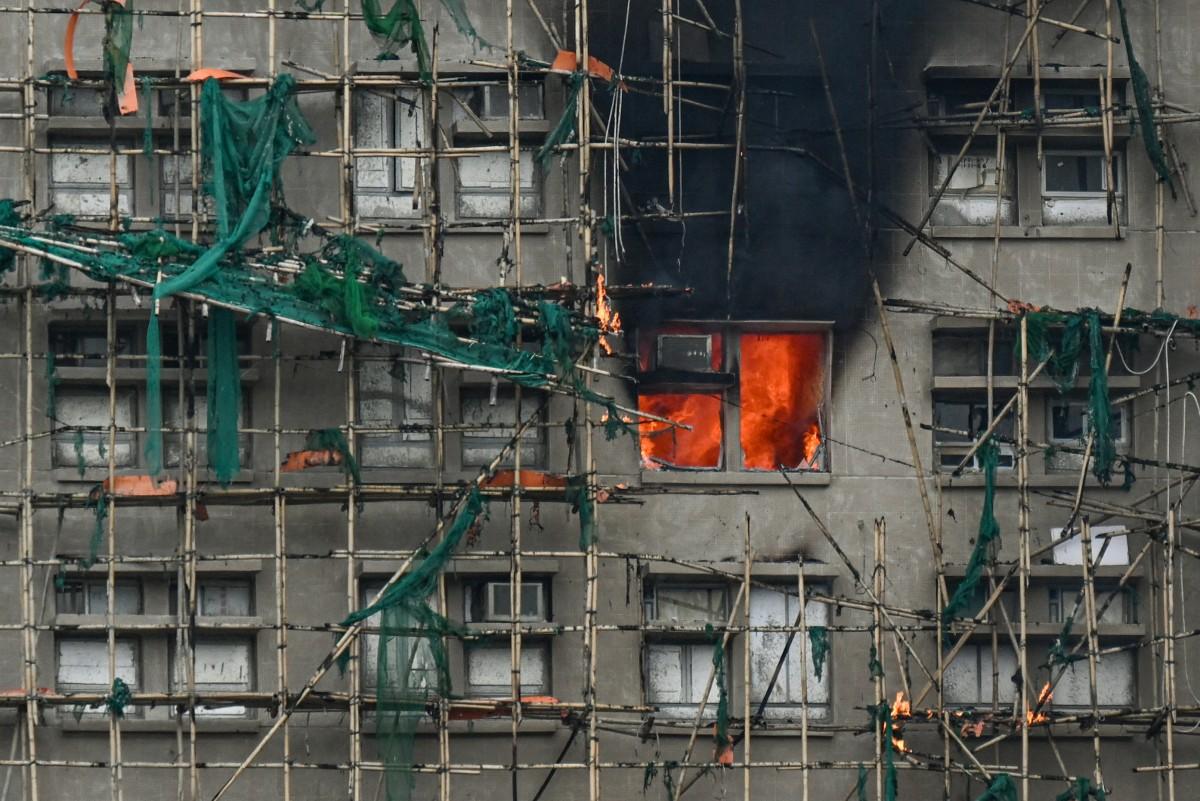 Uma vista de perto de um edifício em construção ou renovação com andaimes de bambu e redes de segurança verde, possivelmente em Hong Kong. Uma janela no centro da estrutura está em chamas intensas, com uma coluna de fumaça preta subindo. O fogo irradia uma luz laranja brilhante para fora da janela.