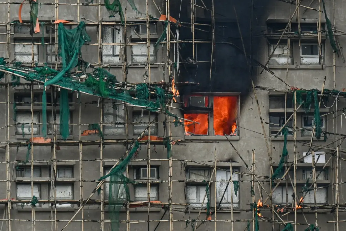 Uma vista de perto de um edifício em construção ou renovação com andaimes de bambu e redes de segurança verde, possivelmente em Hong Kong. Uma janela no centro da estrutura está em chamas intensas, com uma coluna de fumaça preta subindo. O fogo irradia uma luz laranja brilhante para fora da janela.