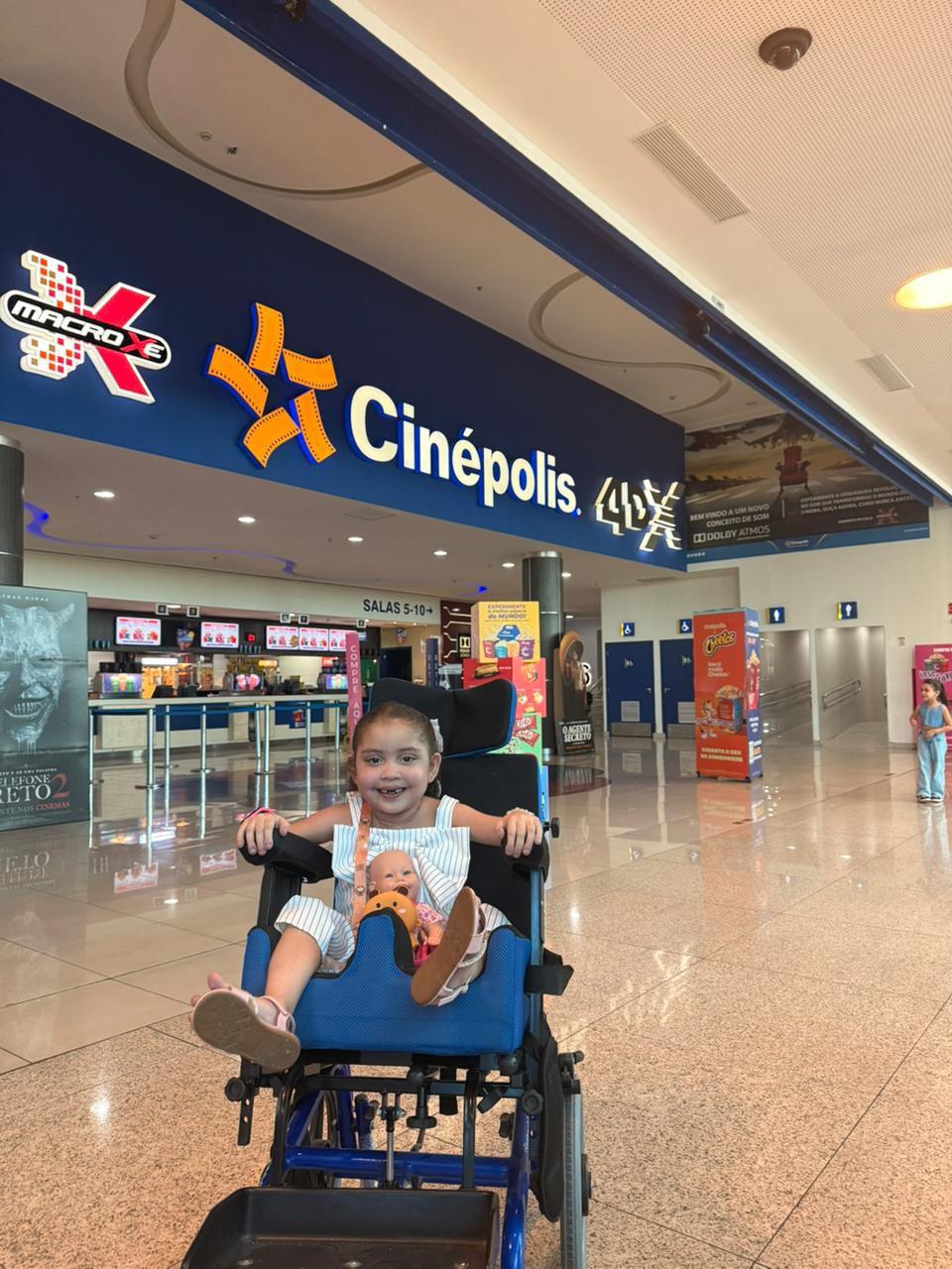 Menina sorridente com feições fofas sentada em carrinho de criança no interior de cinema, em frente ao letreiro do cinema, com ambiente agradável e diversão garantida.