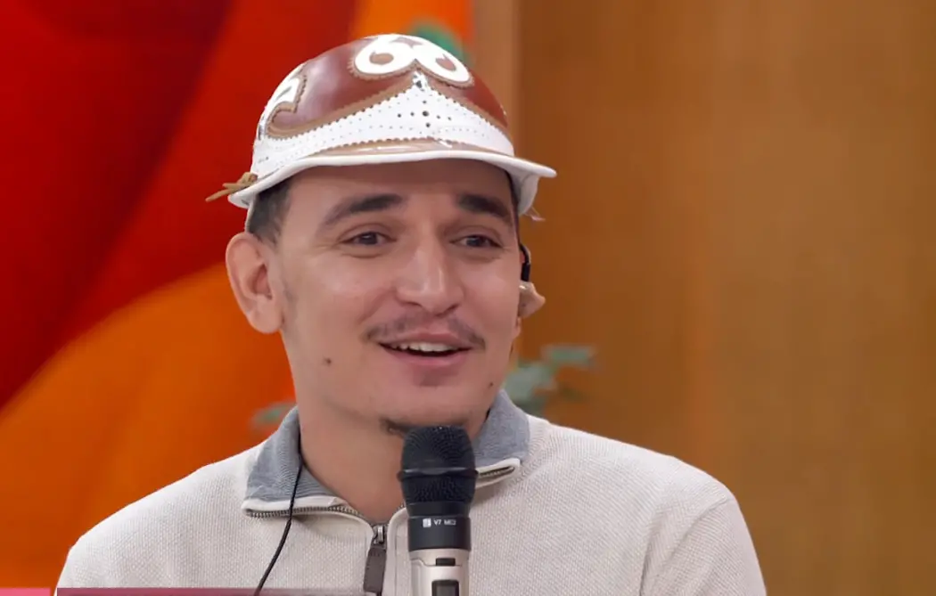 O cantor João Gomes, sorridente, usa um chapéu de couro branco e marrom, típico do Nordeste brasileiro, enquanto segura um microfone em um programa de televisão (Encontro). Ele está vestindo um casaco de moletom cinza claro com gola polo. O fundo é desfocado em tons de laranja e marrom.