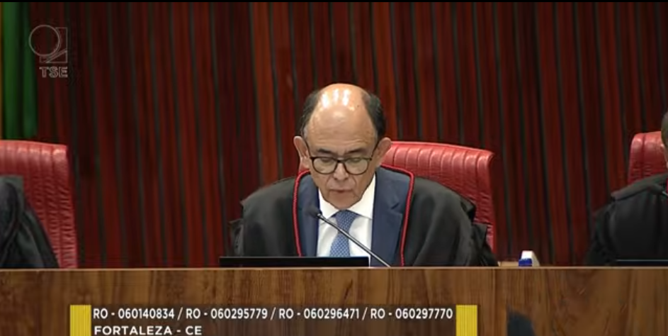 Homem de idade madura, usando toga preta com detalhes em vermelho e camisa com gravata azul, está sentado em uma cadeira alta estofada em vermelho, falando ao microfone durante uma sessão de tribunal. Ao fundo, vê-se uma parede de madeira vertical. Na parte inferior da imagem, aparece uma faixa com informações de processos e a indicação “Fortaleza – CE”.