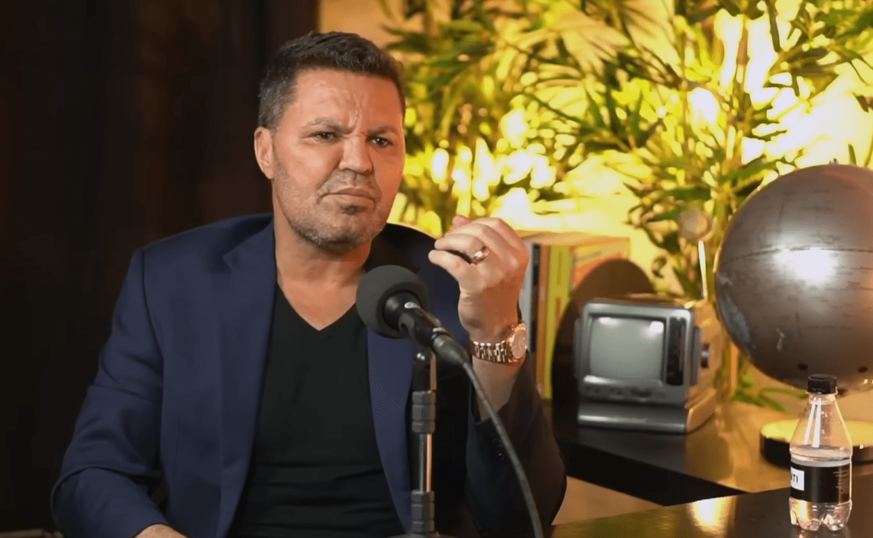 O cantor Eduardo Costa vestindo um blazer azul escuro sobre uma camisa preta, está sentado em uma mesa de estúdio, falando em um microfone de braço ajustável. Ele tem cabelo castanho escuro e uma expressão facial séria enquanto gesticula com a mão direita, que tem um anel e um relógio de pulso dourados. Ao fundo, à direita, há um globo terrestre de metal, uma garrafa de água e um pequeno monitor de televisão antigo, com uma iluminação amarela quente de plantas desfocadas atrás. A cena parece ser de um podcast ou entrevista.