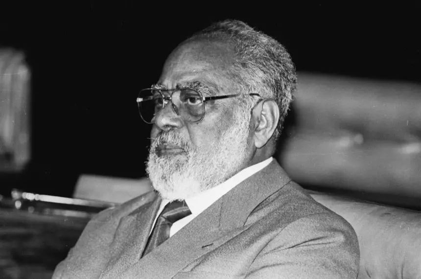 Uma foto em preto e branco de Abdias Nascimento, político, artista e ativista do movimento negro brasileiro. Ele aparece em um plano de médio-close-up, de perfil, vestindo um terno e gravata, com barba e óculos. Ele olha pensativo para a esquerda.