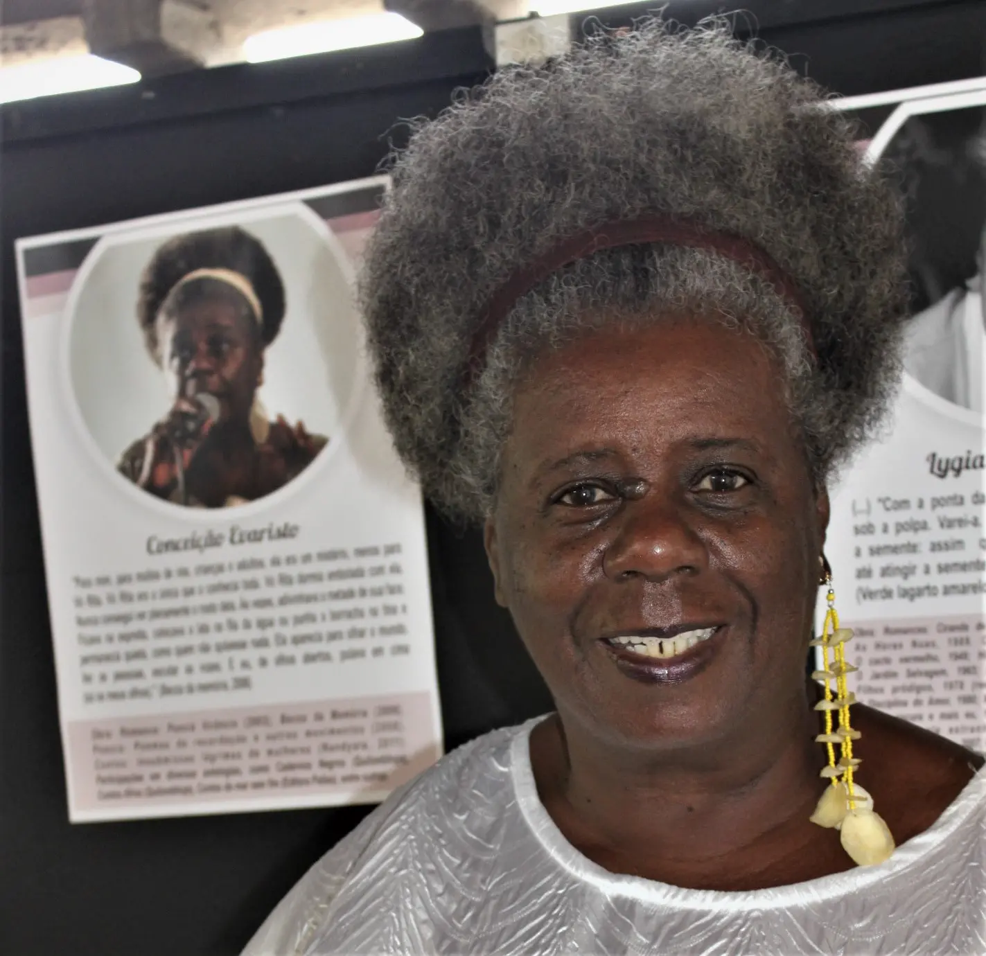 Um close-up da escritora Conceição Evaristo, sorrindo para a câmera. Ela usa uma roupa branca e uma tiara escura em seu cabelo crespo grisalho. Ao lado, uma placa exibe uma foto e uma citação sua.