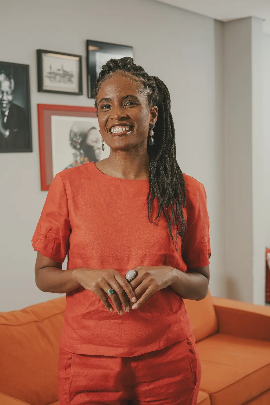 Um retrato em cores da filósofa e ativista Djamila Ribeiro, vestida com um conjunto de calça e blusa cor de telha. Ela sorri para a câmera, com tranças longas e brincos em evidência, em um ambiente interno com quadros pendurados na parede atrás dela.