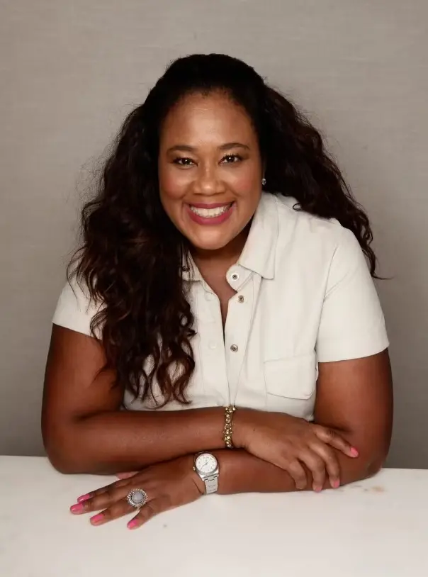 Um retrato em cores da dermatologista Katleen Cruz. Ela tem longos cabelos escuros, veste uma camisa clara de colarinho e sorri. Ela está sentada em uma mesa, com relógio e anéis em destaque.