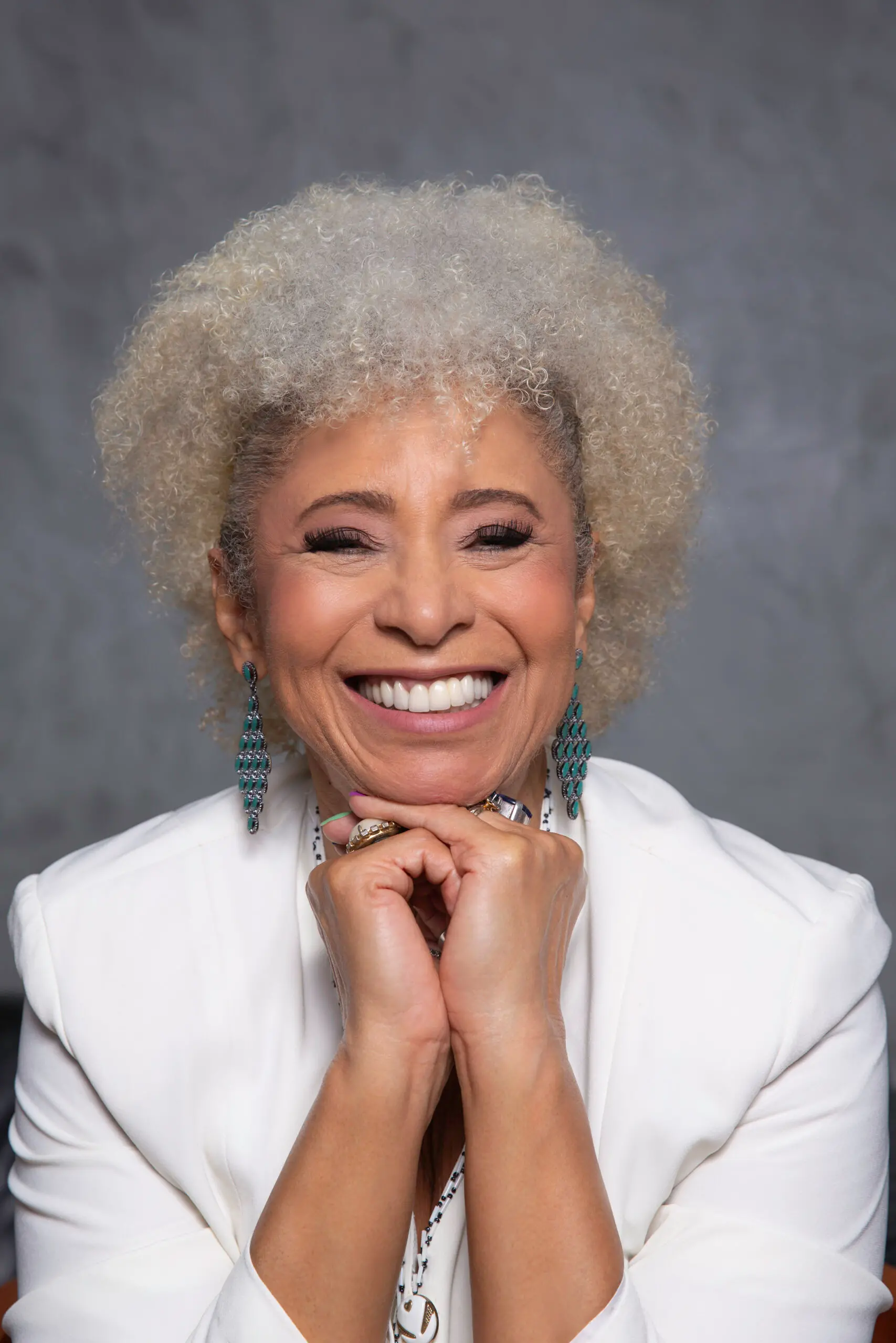 Um retrato em cores da física Sonia Guimarães, a primeira mulher negra brasileira doutora em Física. Ela tem cabelos crespos loiros/grisalhos e veste um blazer branco, sorrindo abertamente, com as mãos sob o queixo e brincos turquesa longos.