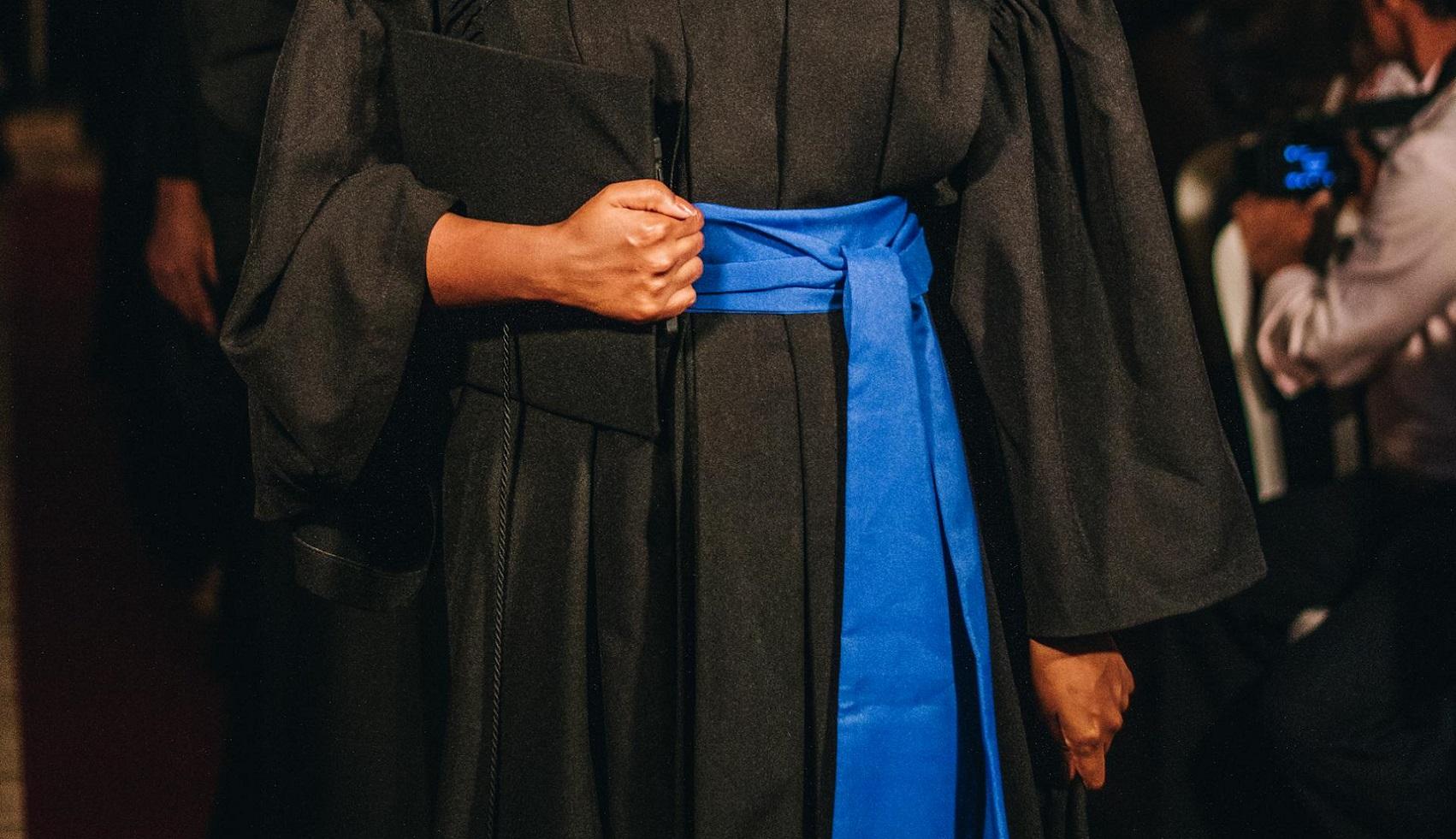 Foto em close-up de uma mulher negra vestida de toga preta de formatura, com faixa azul enrolada na cintura. Ela segura uma pasta preta junta ao corpo. Atrás dela, há uma fila de mais pessoas formadas e, ao lado, há um fotógrafo.